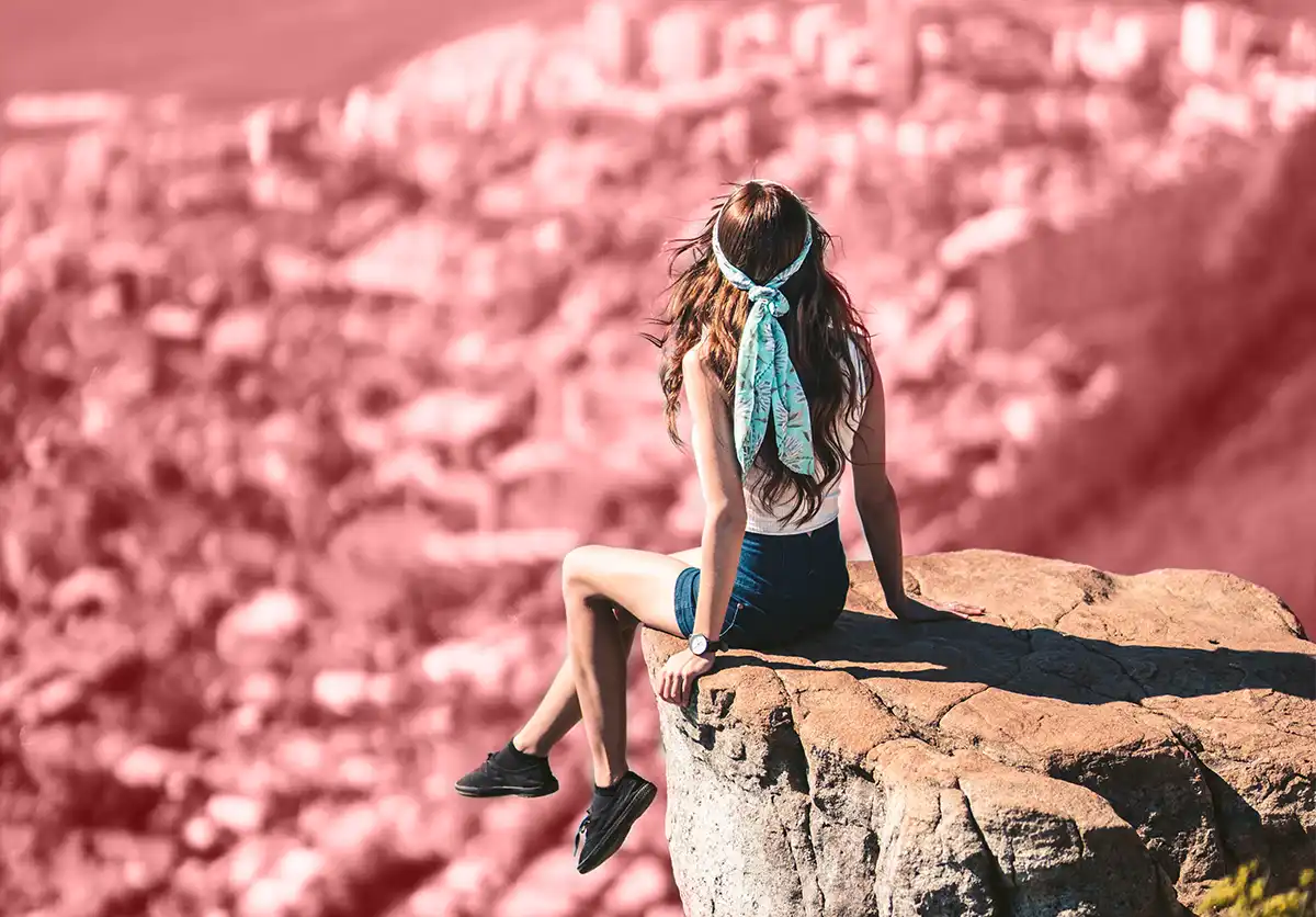 Mujer con pañuelo celeste en el pelo, sentada en el borde de un acantilado, observando una ciudad a lo lejos.